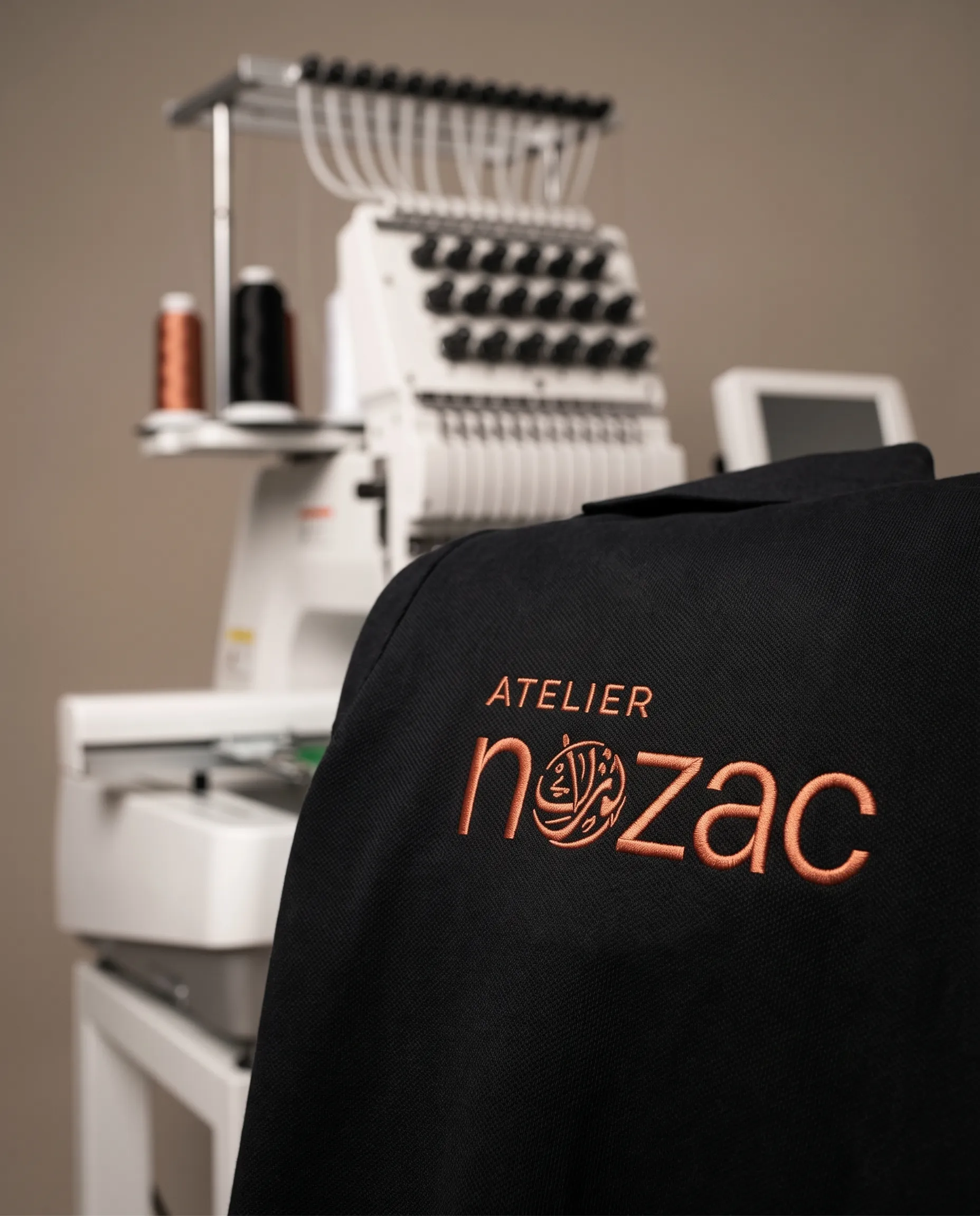 Logo Nozac brodé en cuivre sur textile noir, machine à broder en arrière-plan
